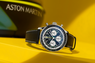 Die Breitling Navitimer B01 Chronograph Aston Martin Edition ist schräg auf einem gelben Podest inszeniert, im Hintergrund ist unscharf das Aston-Martin-Branding zu erkennen.