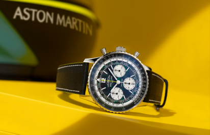 Die Breitling Navitimer B01 Chronograph Aston Martin Edition ist schräg auf einem gelben Podest inszeniert, im Hintergrund ist unscharf das Aston-Martin-Branding zu erkennen.