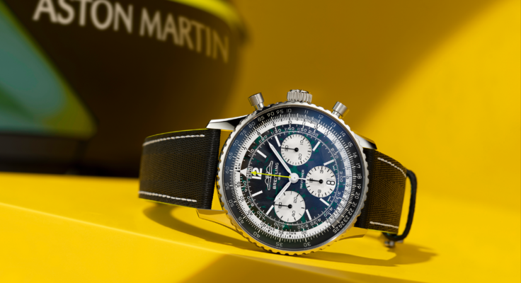 Die Breitling Navitimer B01 Chronograph Aston Martin Edition ist schräg auf einem gelben Podest inszeniert, im Hintergrund ist unscharf das Aston-Martin-Branding zu erkennen.